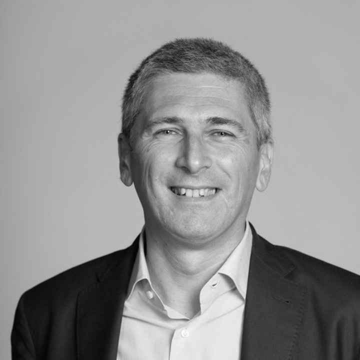 Black-and-white portrait of a smiling, short-haired person wearing a jacket and collared shirt, posed against a simple background, embodying the confidence needed to navigate strategic sourcing in an ever-changing supply chain landscape.