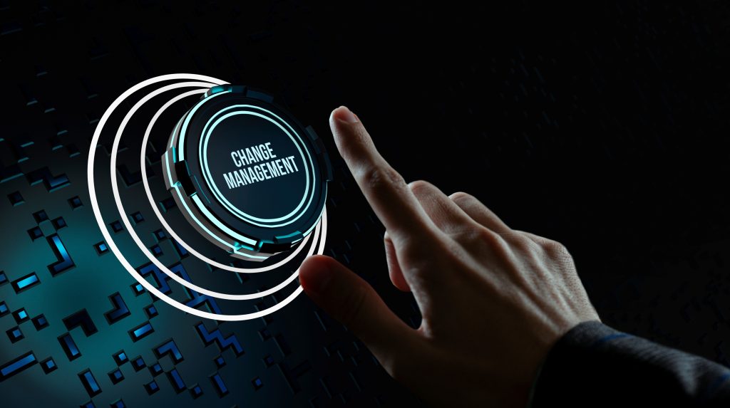 Una mano si allunga verso un pulsante luminoso con la scritta "Change Management", incastonato in uno sfondo digitale oscuro. Anelli illuminati circondano il pulsante, evocando un'interfaccia virtuale high-tech che allude a transizioni fluide e innovazione.