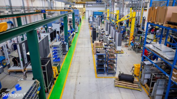 L'ampio interno della fabbrica, sede di SWEGON OPERATIONS, è caratterizzato da travi strutturali blu e verdi. Durante il tour della fabbrica, vedrai i lavoratori diligentemente alle loro postazioni organizzate lungo le linee di montaggio sotto soffitti alti in questo spazio ben illuminato.