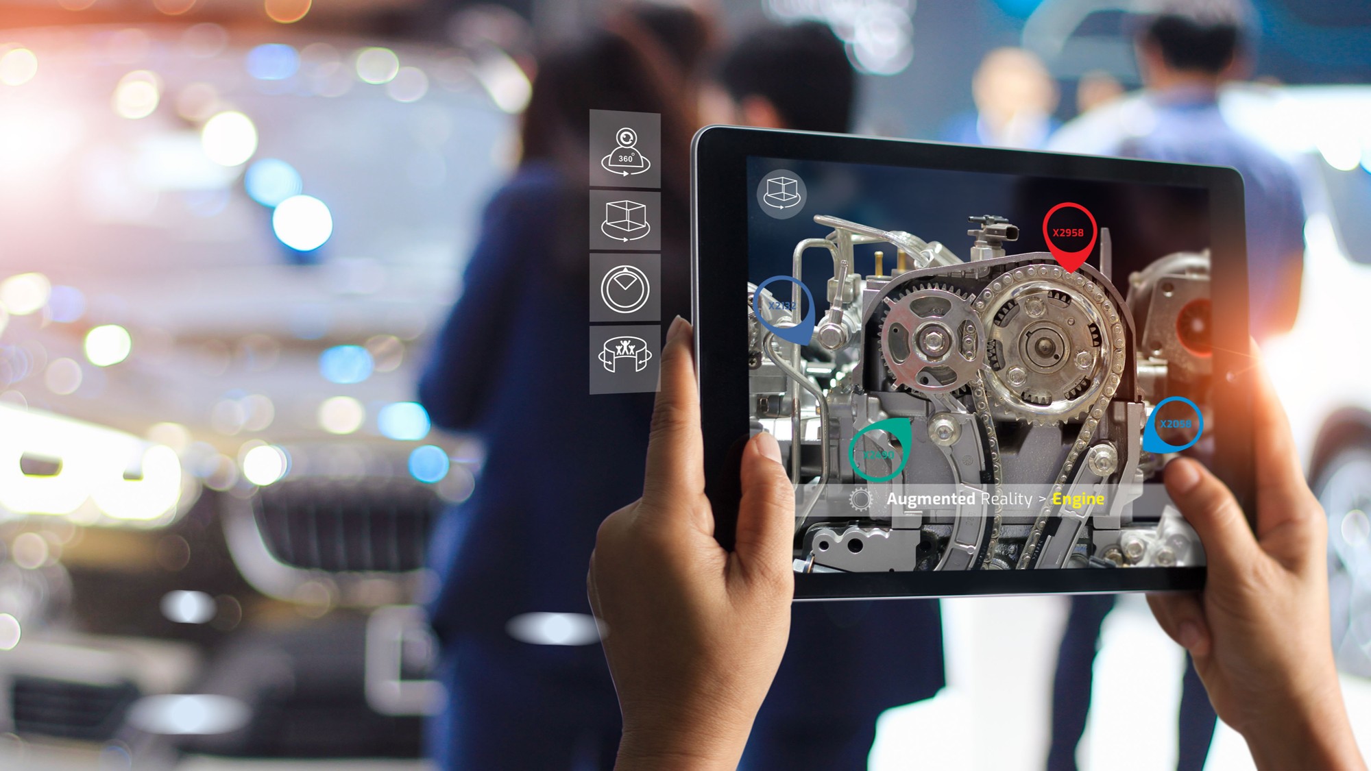 Una persona tiene in mano un tablet che mostra una sovrapposizione di realtà aumentata di parti del motore e diagnostica, illustrando un sistema di formazione sulla produzione in una fiera automobilistica sfocata con persone e un'auto sullo sfondo.