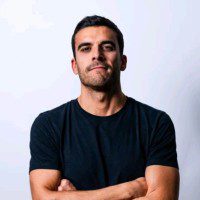 Un uomo con capelli corti e scuri e barba curata si staglia su uno sfondo chiaro, con le braccia incrociate, indossando una semplice maglietta nera e guardando con sicurezza verso la telecamera, incarnando l'innovazione e l'impatto misurabile dell'IA industriale.