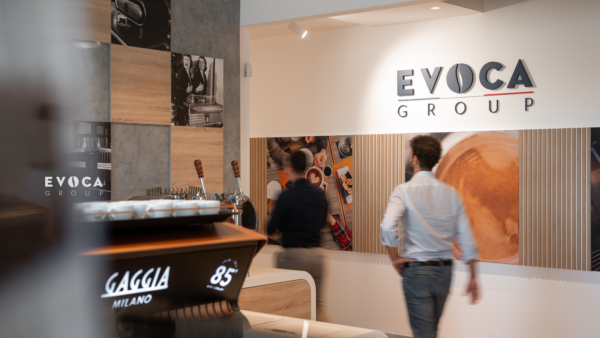 Due persone attraversano un ufficio o uno showroom moderno con le insegne di Evoca Group sulla parete. Una macchina da caffè Gaggia Milano è in primo piano, mentre l'eccellenza operativa e l'arredamento a tema caffè sono sullo sfondo.