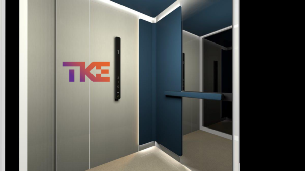 Modern elevator interior with sleek metallic walls, a blue accent panel, a mirror, and a horizontal handrail – ideal for factory tour routes or supply chain management-focused sites. The TK Elevator TKE logo is prominently displayed on the wall.