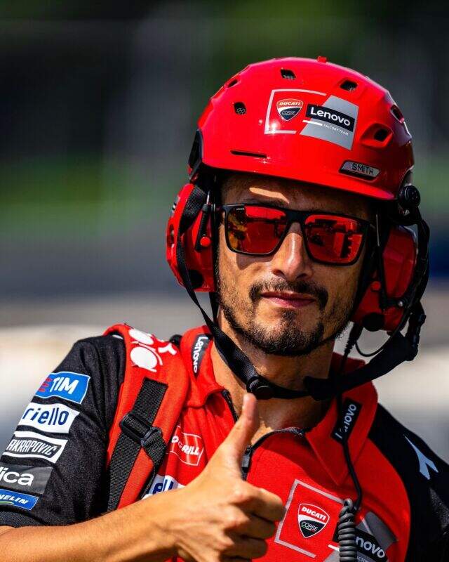 Un uomo che indossa un casco rosso, occhiali da sole rossi e una tenuta da corsa Ducati Lenovo fa il gesto del pollice in su, in piedi all'aperto sotto la luce del sole.
