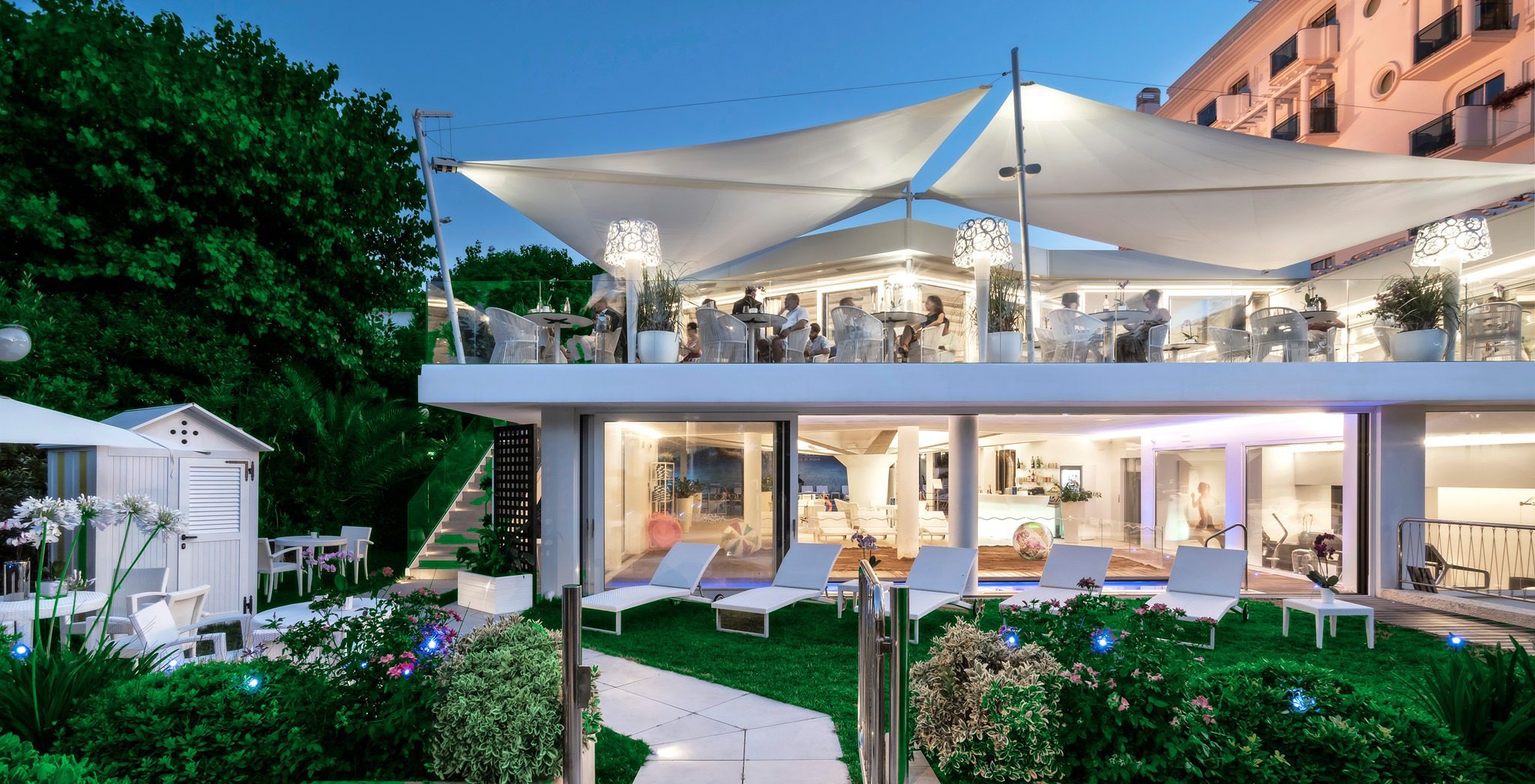 Un moderno hotel a più piani con balconi e una zona pranzo sul tetto, illuminato al tramonto sotto un cielo blu con nuvole sparse. In primo piano, un paesaggio lussureggiante e posti a sedere all'aperto.