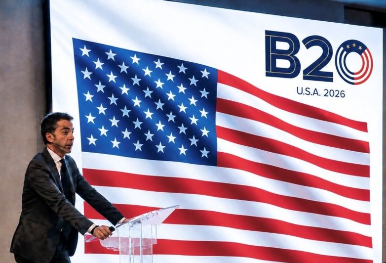 A man in a suit and tie speaks from a podium in front of a large backdrop with the American flag and the words “B20 U.S.A. 2026,” addressing key themes related to B20 economic growth within the broader G20 agenda.